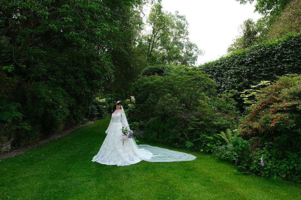 A stunning bride stands elegantly on a lush green lawn surrounded by mature trees and rich garden foliage, holding a bouquet and wearing a lace gown with an off-the-shoulder neckline and a long flowing veil.