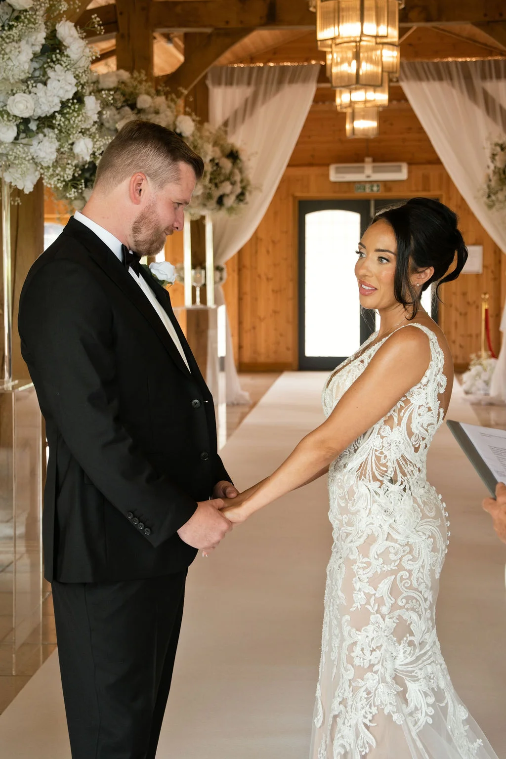 The bride and groom hold hands and exchange loving glances during their indoor wedding ceremony inside an elegant wood-beamed barn venue.
