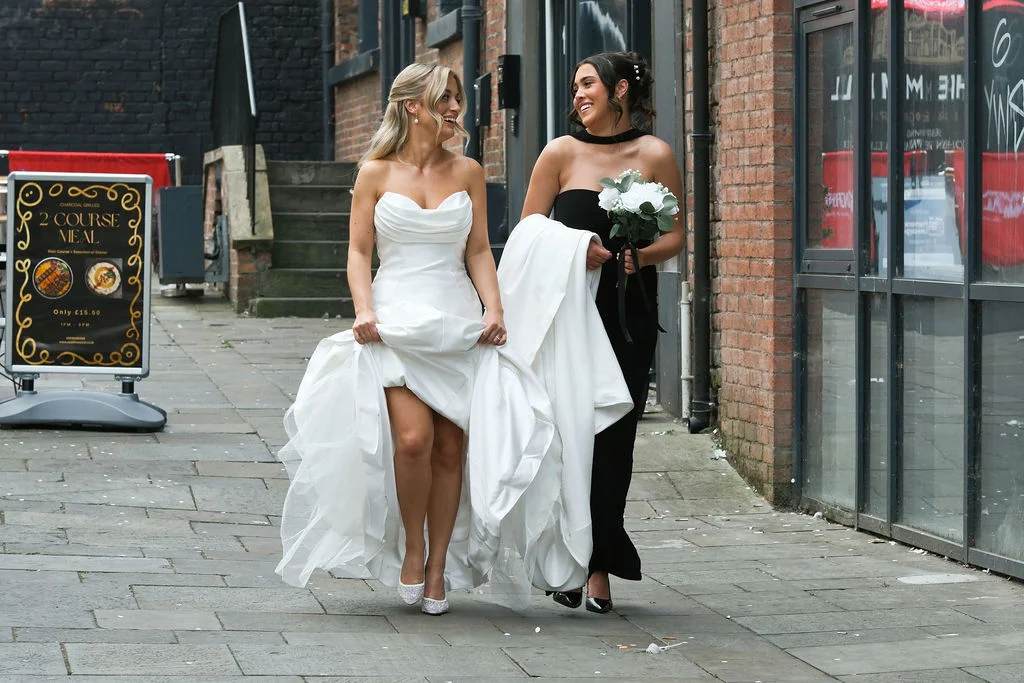 A bride in a strapless white gown and her bridesmaid in a black dress walk and laugh together down a brick-lined city street.
