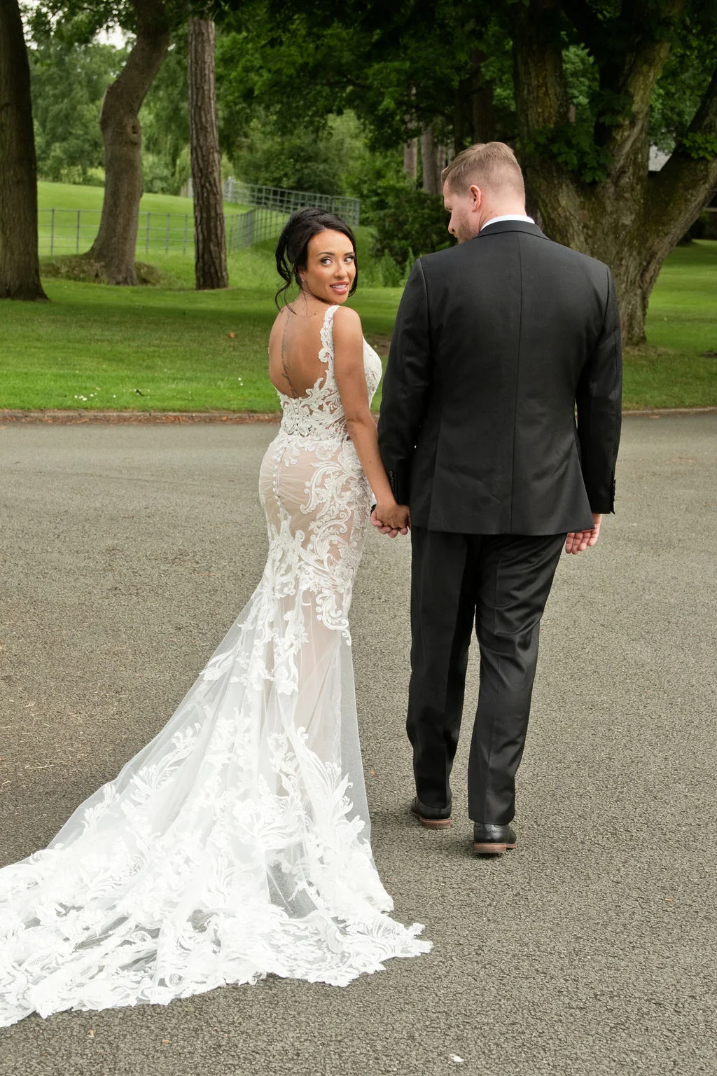 A newlywed couple walking hand-in-hand along an outdoor path, with the bride looking back over her shoulder wearing a lace mermaid gown with a dramatic train and open back.