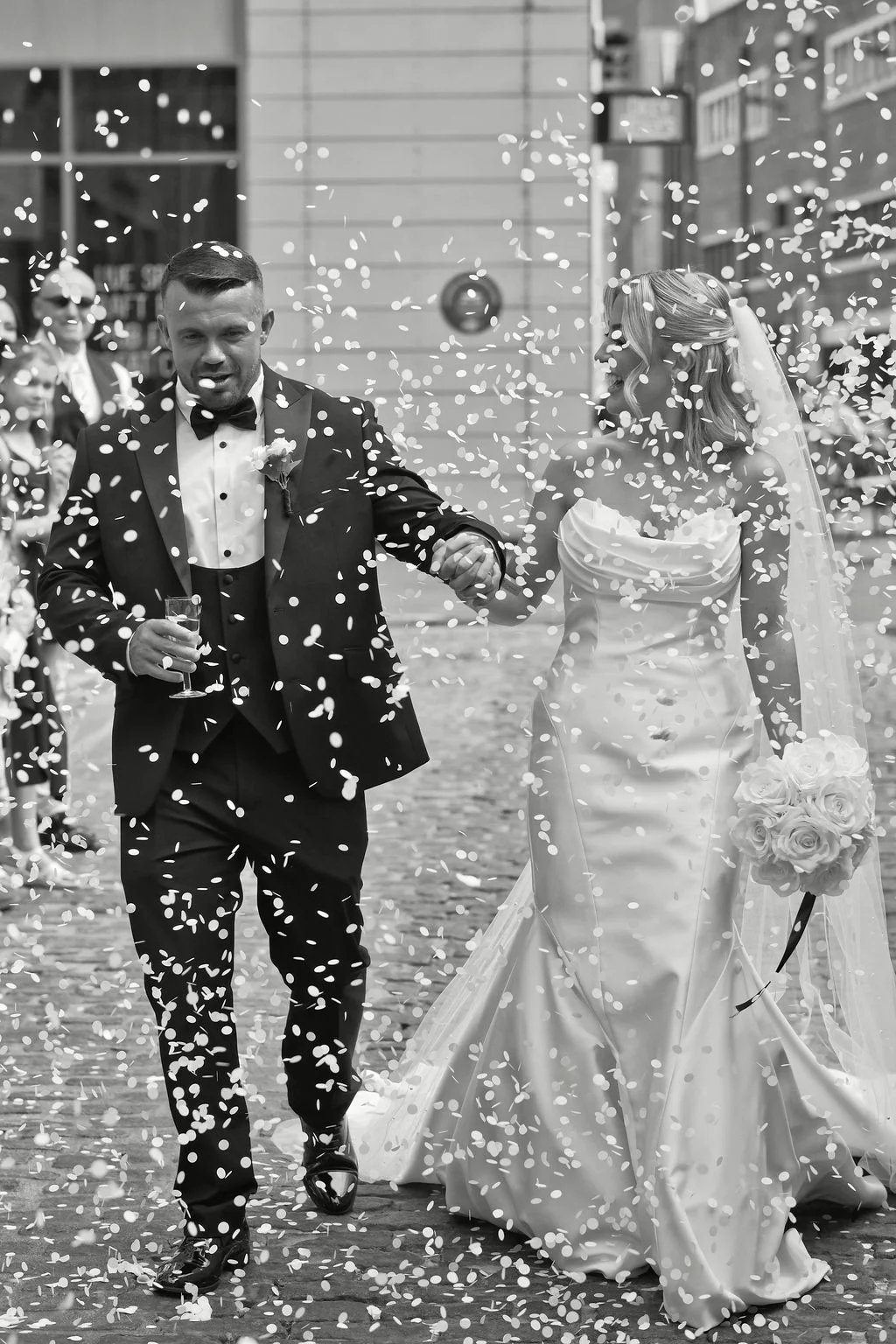 A joyful newlywed couple walks hand in hand down a cobbled city street amidst a cascade of falling confetti during their wedding celebration.