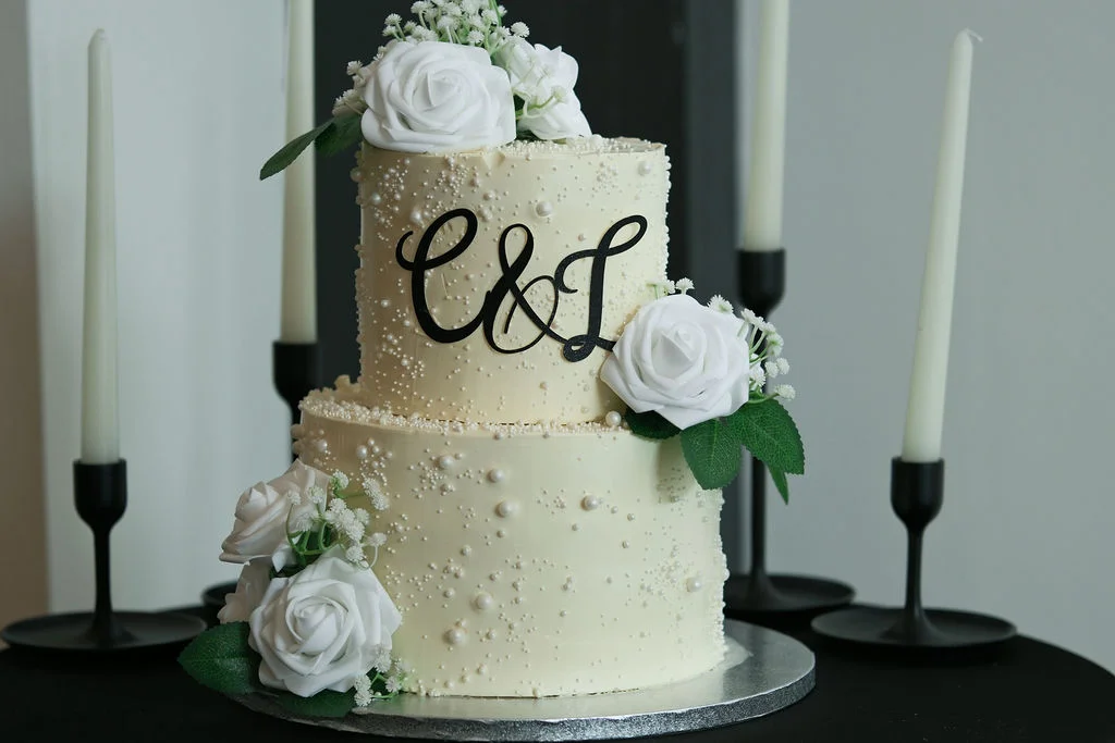An elegant two-tier wedding cake decorated with pearl accents and white roses sits on a table flanked by tall black candlesticks.