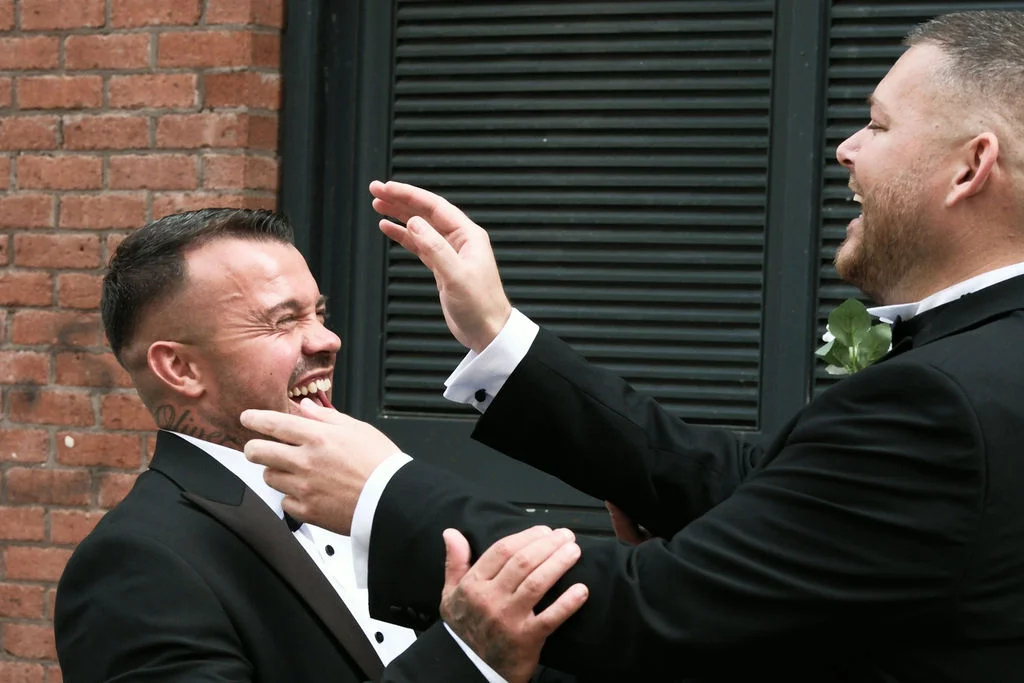 Two grooms share a joyful belly laugh together outdoors against an exposed brick wall, wearing smart black tuxedos with bow ties.