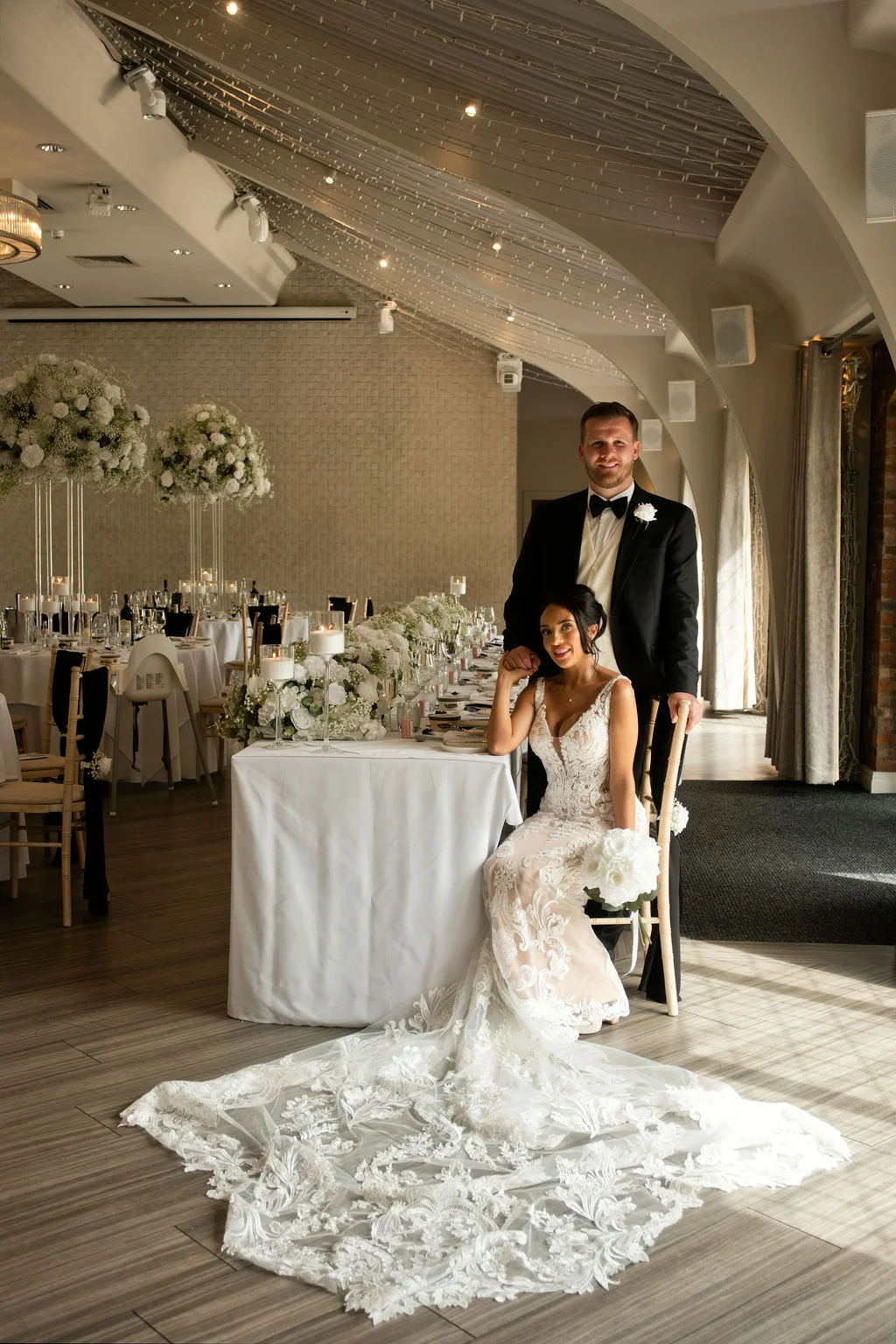 A newlywed couple poses together at a beautifully decorated top table inside a modern venue featuring a dramatic lace train on the bride's gown.