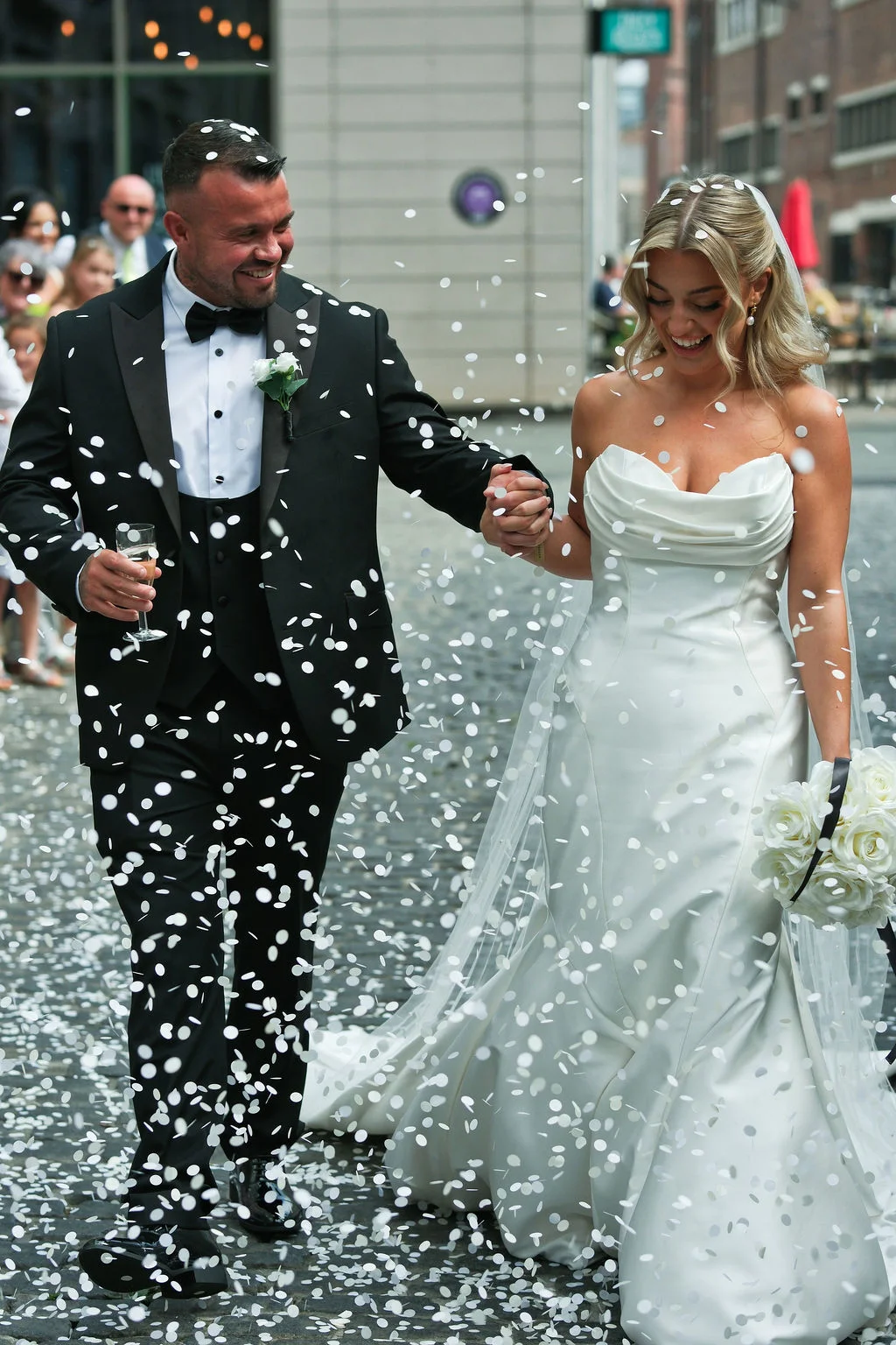 A joyful newlywed couple laughs happily as they walk hand-in-hand through a flurry of white confetti on a city street.