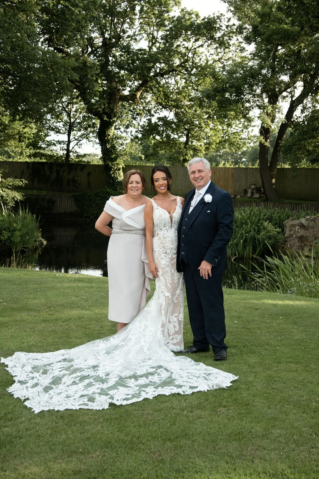A smiling bride in an ornate lace gown with a long train poses outdoors on a lawn beside her parents near a pond on her wedding day.