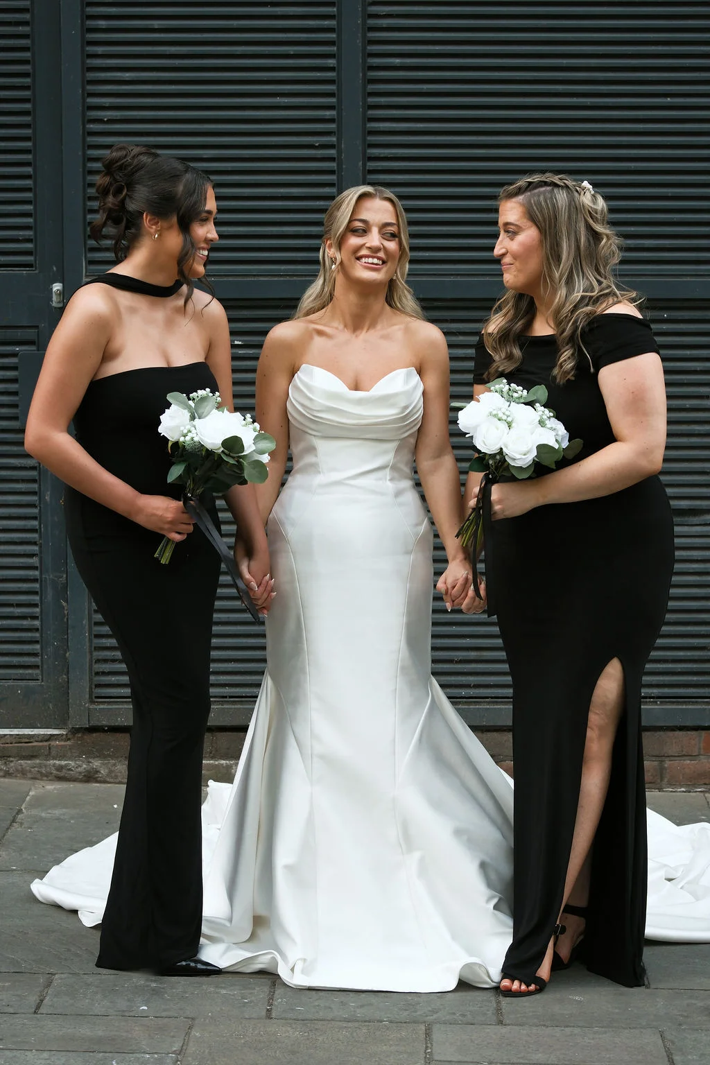 A radiant bride in a strapless white satin gown stands smiling while holding hands with her two bridesmaids wearing elegant black dresses and holding white rose bouquets.