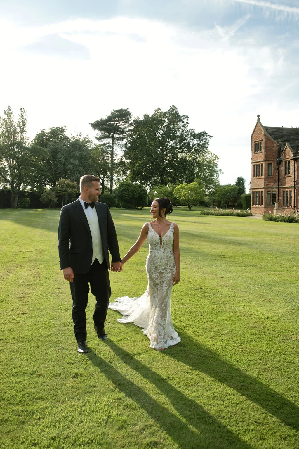 Newlyweds smiling at each other while walking hand in hand across a manicured lawn in front of a grand country house.