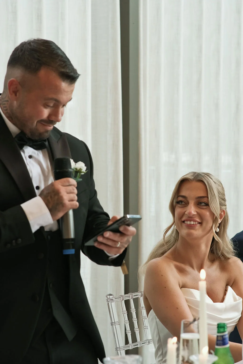 A smiling groom delivers his wedding speech reading from a smartphone while his bride looks on lovingly at the reception table.