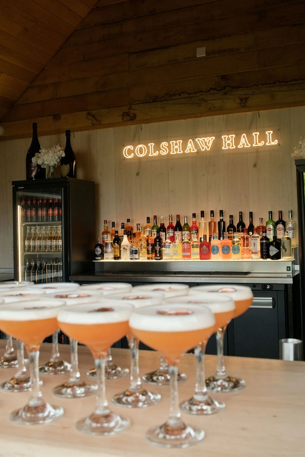 A well-stocked wooden wedding bar features rows of colourful cocktails in coupe glasses in the foreground beneath a glowing custom neon sign.