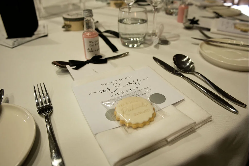 A close-up view of an elegantly set wedding table featuring a white tablecloth, personalised stationery with a custom cookie favour, and sleek black cutlery.