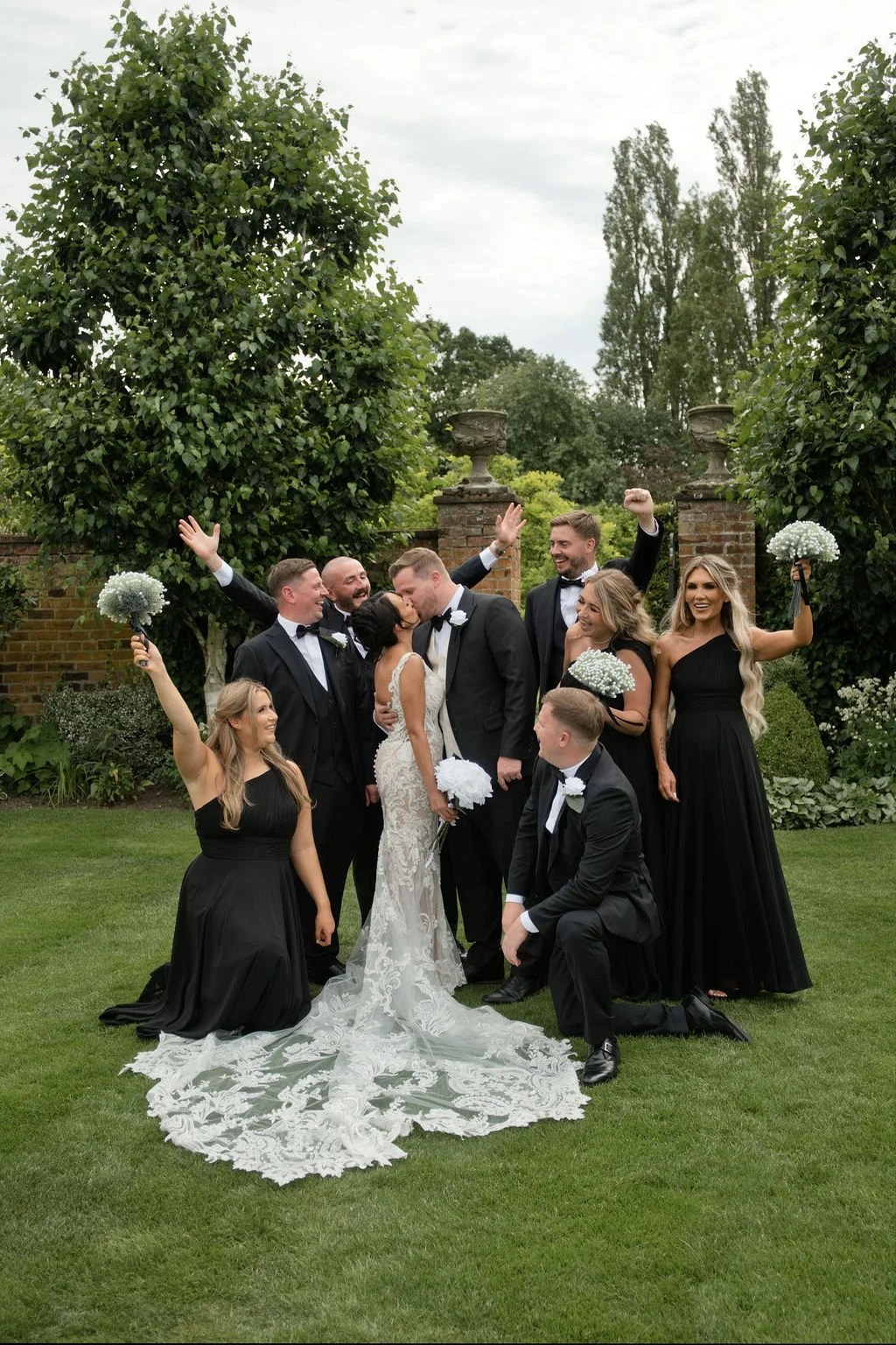 The bride and groom share a kiss surrounded by their wedding party in black attire on a lush green lawn.