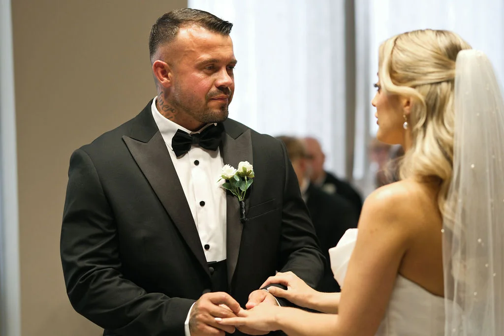 A groom wearing a black tuxedo and bow tie places a ring on his bride's finger during their wedding ceremony.