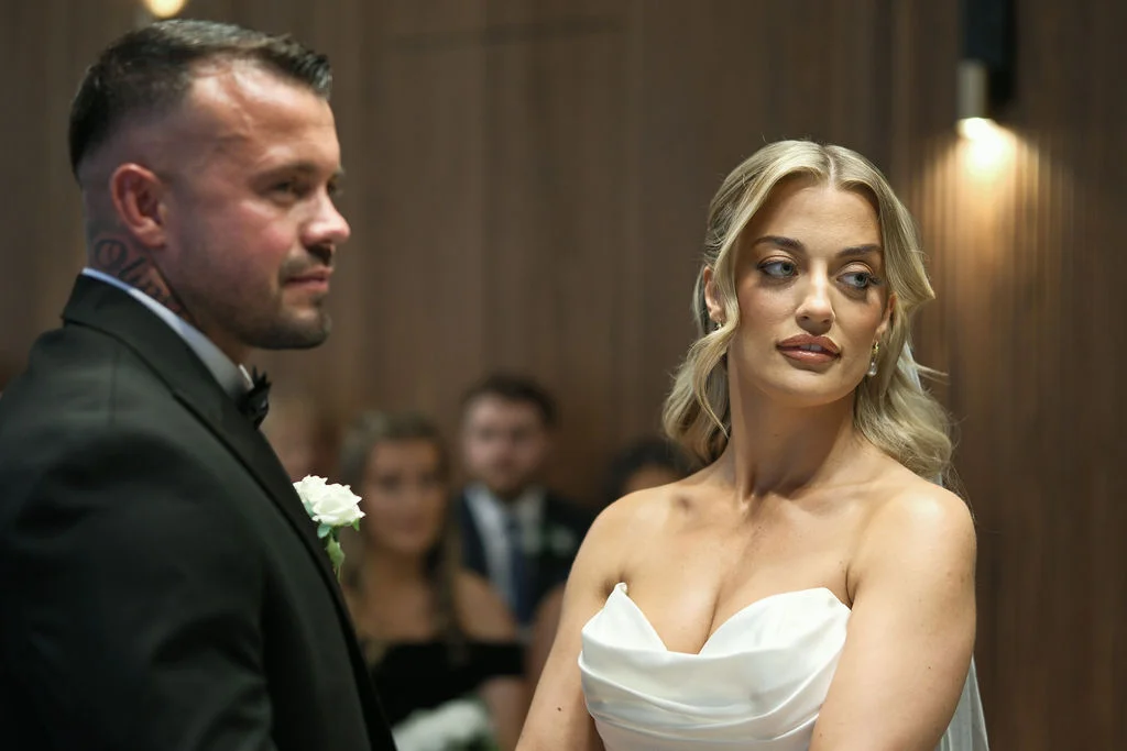 A newly married couple during their civil ceremony, with the bride looking thoughtfully to the side while wearing a sleek strapless satin gown with a cowl neckline.