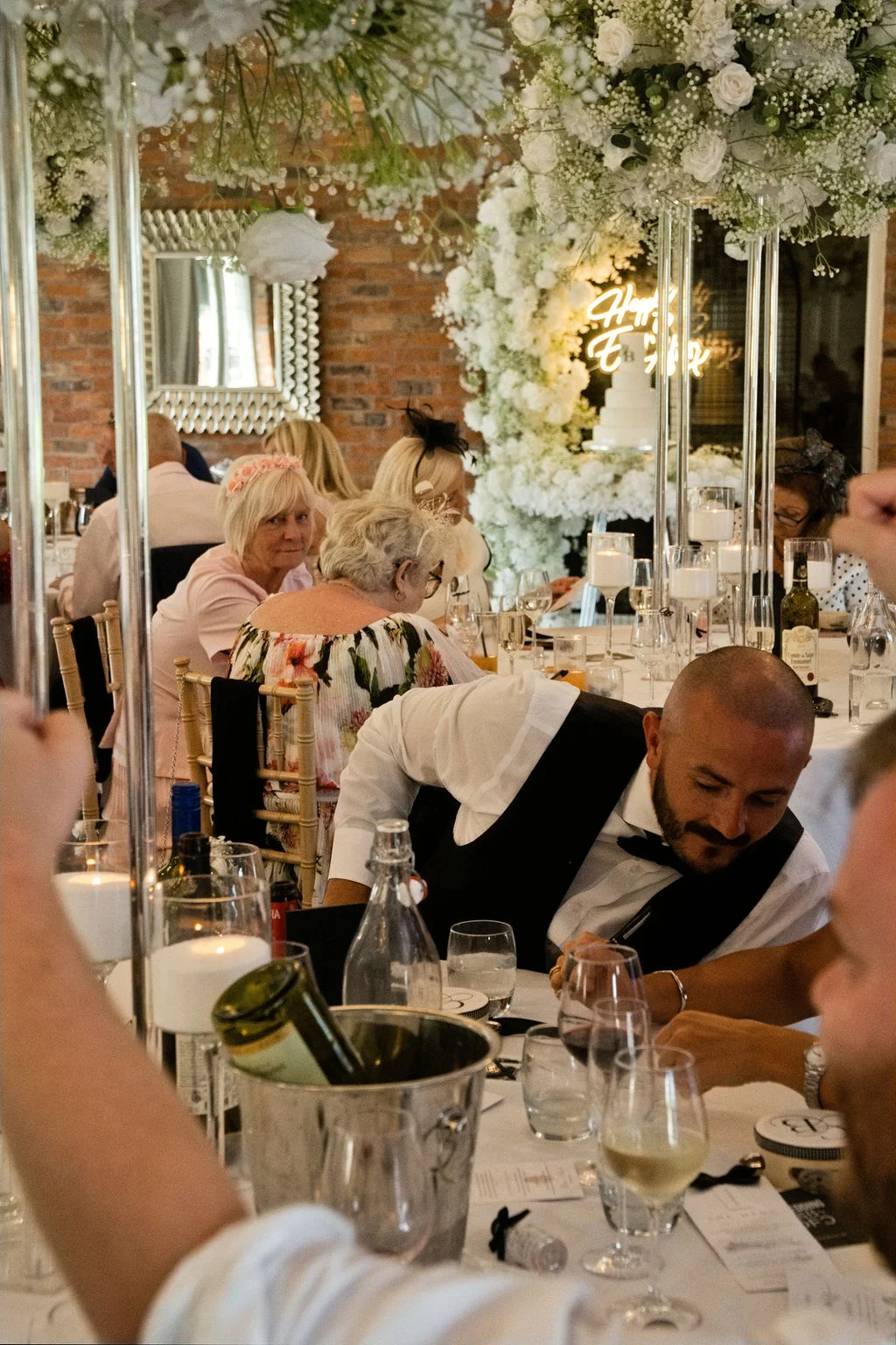 A candid shot of guests seated at round tables during a wedding reception, featuring tall floral centrepieces, a neon sign in the background, and a groom in a waistcoat looking down at the table.
