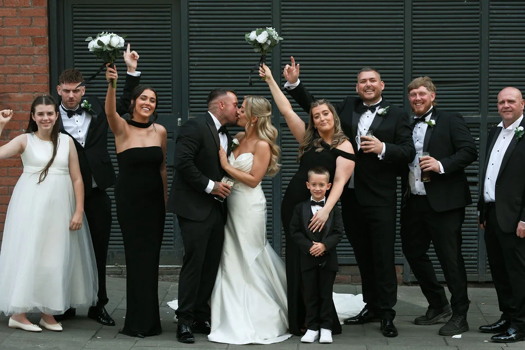 A newlywed couple shares a kiss surrounded by their wedding party dressed in black tuxedos and black bridesmaid dresses outdoors.