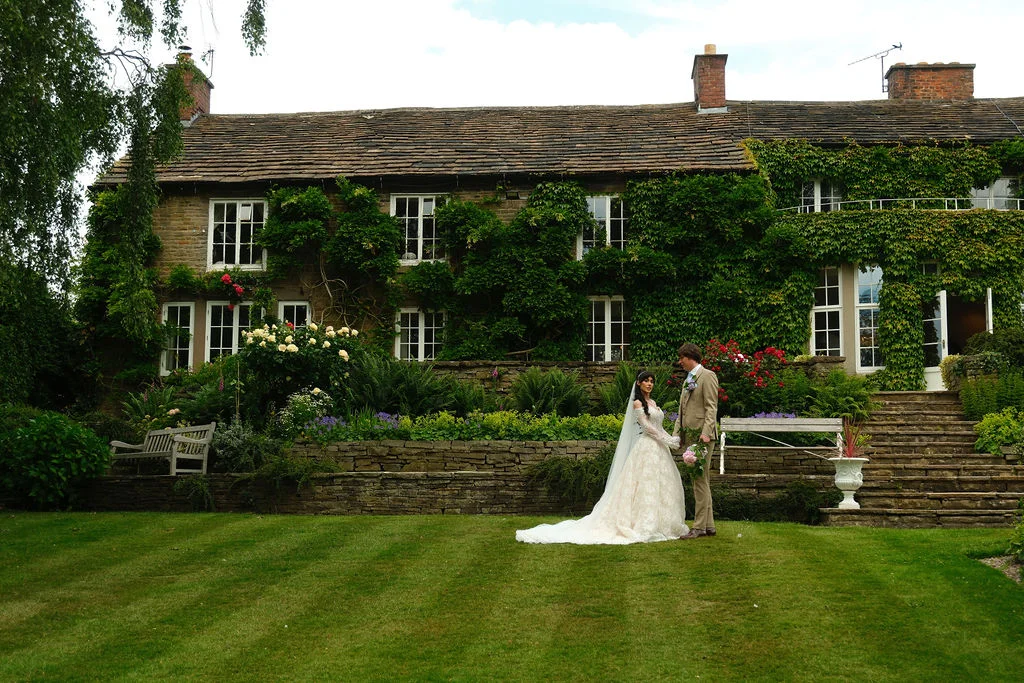 A newlywed couple stands on a lush green lawn in front of a historic stone manor house covered in climbing ivy.