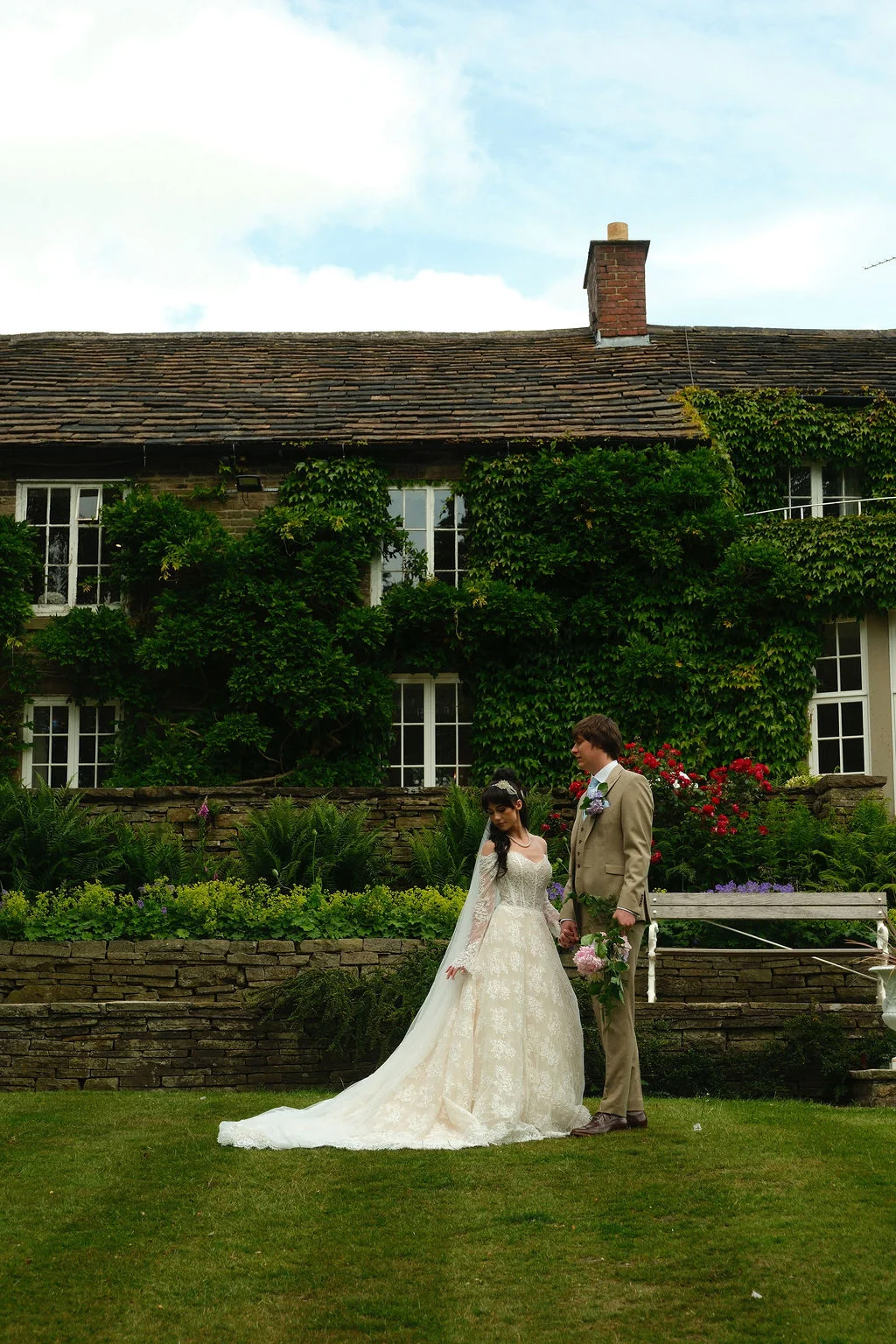 A newlywed couple poses together on a lawn in front of an ivy-clad stone country house, with the bride wearing an intricate lace gown featuring long sleeves and a sweeping train.