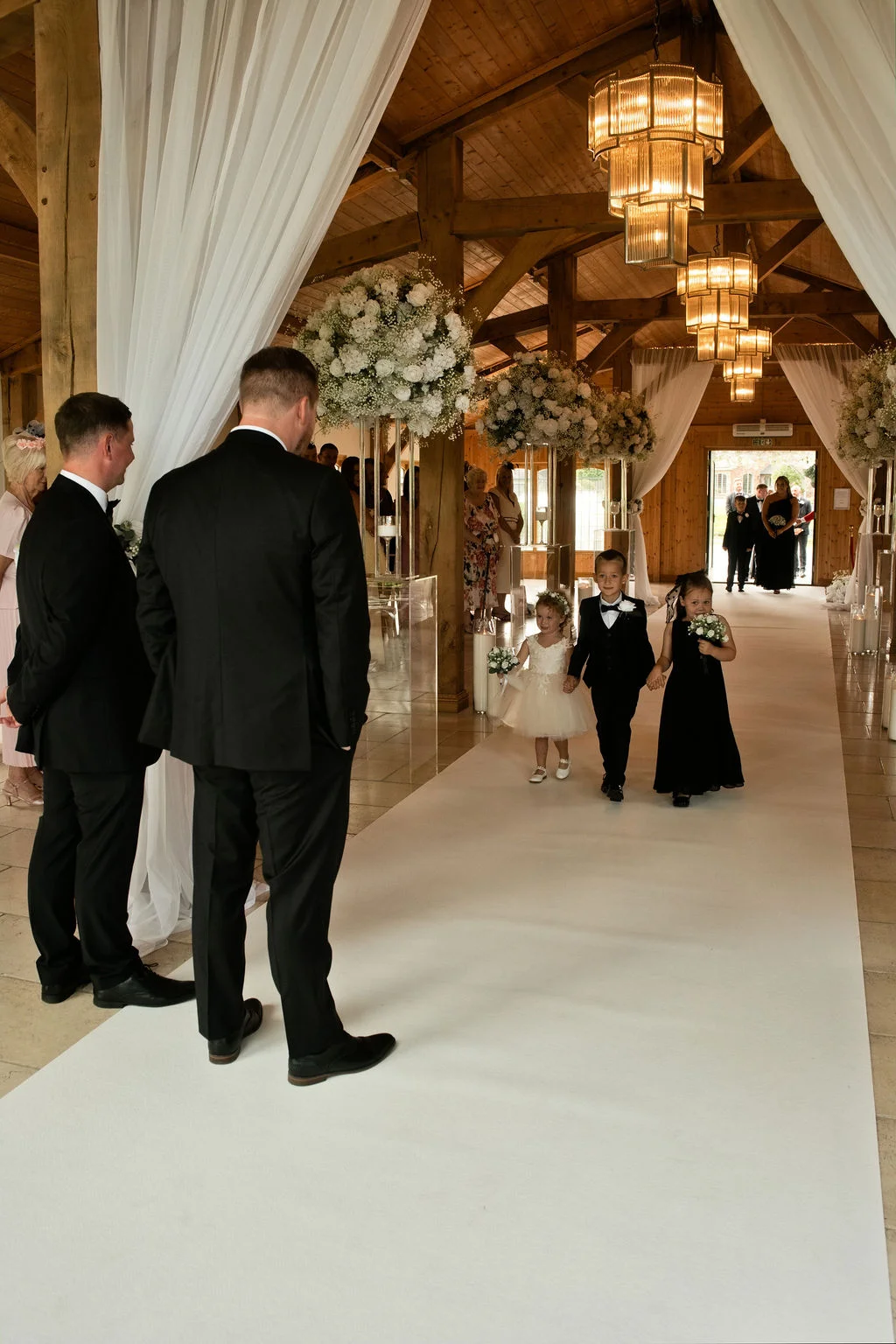A page boy and two flower girls walk down a white aisle runner in a rustic oak-framed barn as groomsmen in black tuxedos look on.