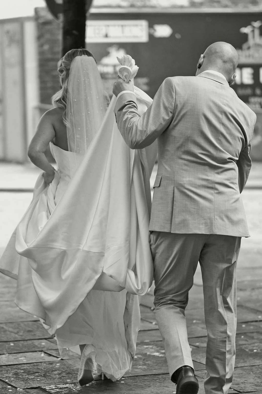 Newlyweds walking away together with the groom holding the bride's satin gown and veil as they navigate a city street.