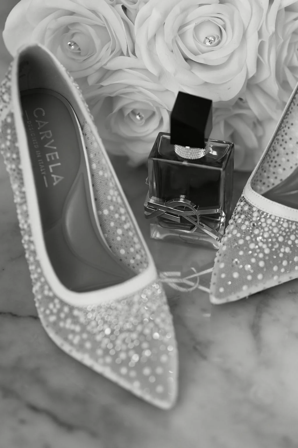 An elegant black and white flat lay showcases sparkly bridal heels alongside a perfume bottle and a bouquet of white roses.