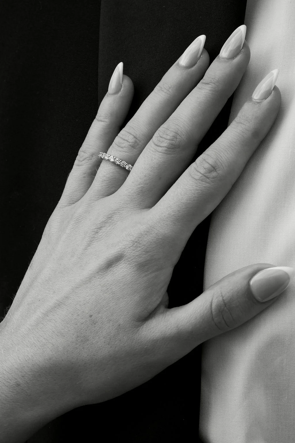 A close-up black and white photograph highlights a hand adorned with a diamond wedding band and French manicure nails against a contrasting dark and light background.