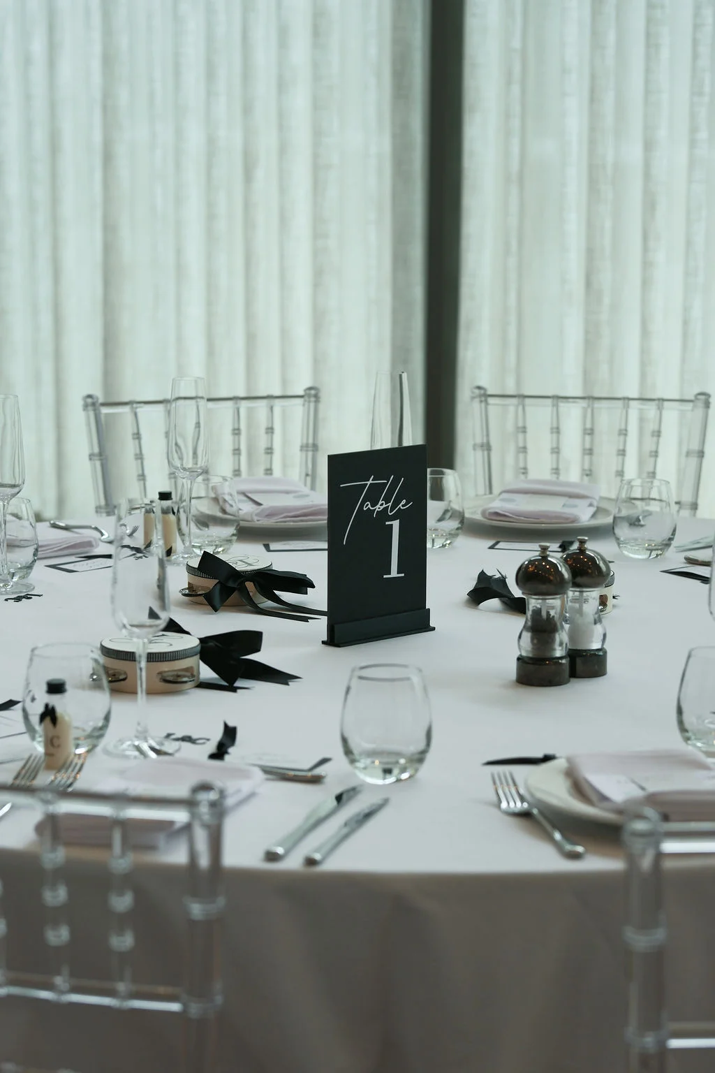 An elegantly styled round wedding table featuring a black table number one card, modern monochrome place settings, glassware, and clear ghost chairs.