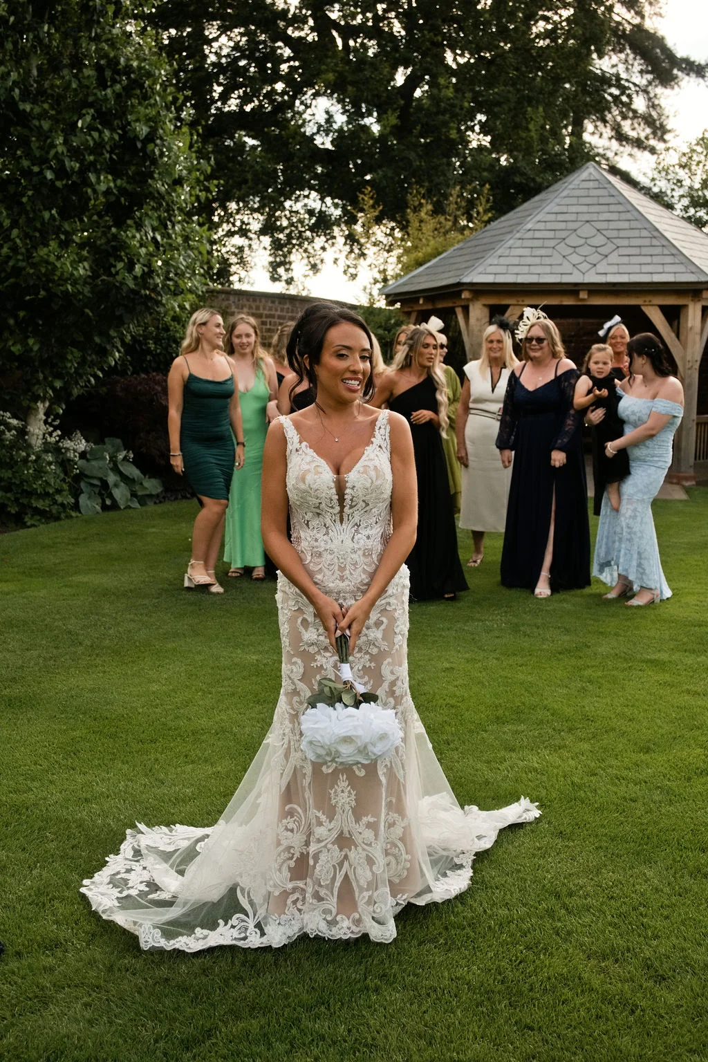 A smiling bride holding a white rose bouquet stands on a manicured lawn in her lace gown while wedding guests look on near a wooden gazebo.