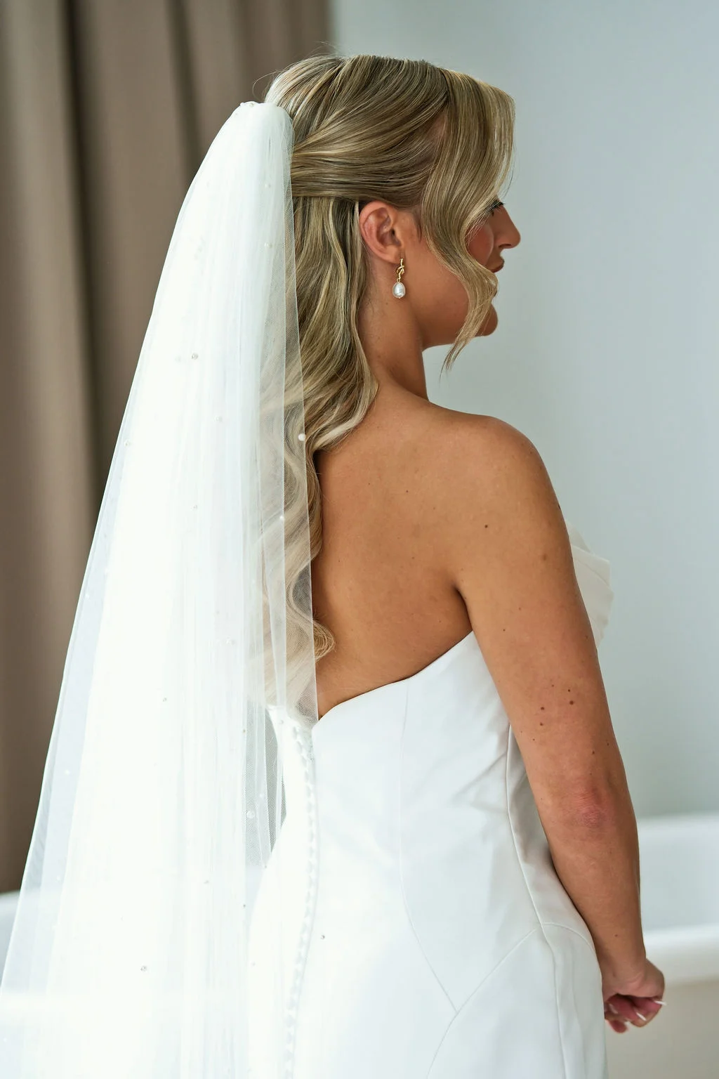 A profile view of a bride with soft waves and a pearl veil, wearing a strapless white wedding gown.