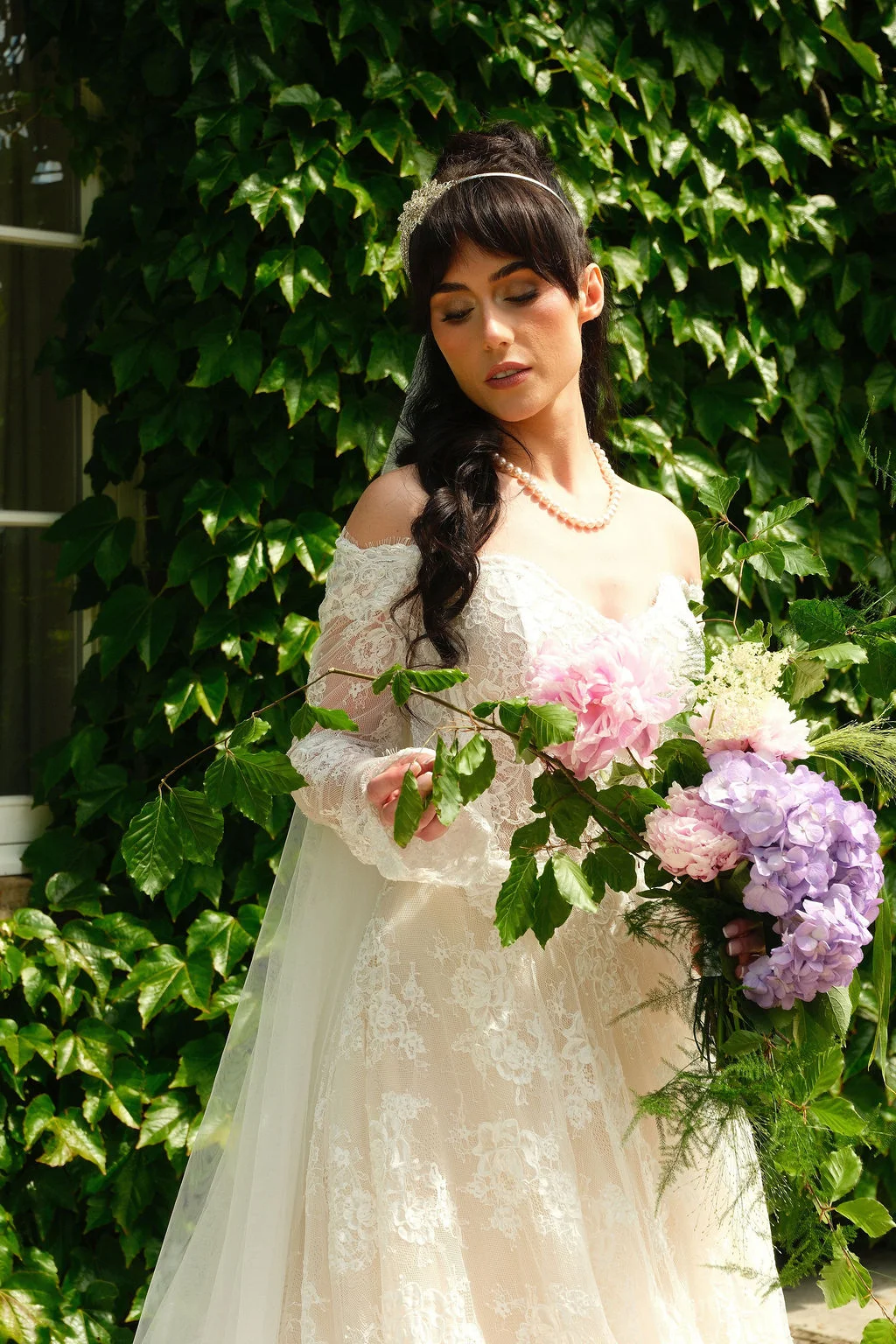 A stunning bride with dark hair and a fringe poses against an ivy-covered wall, wearing an off-the-shoulder lace gown, a pearl necklace, and holding a lush bouquet of pink and purple hydrangeas and peonies.