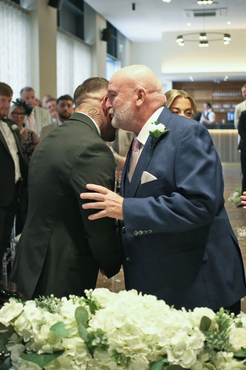 A groom embracing and kissing his partner on the neck during a warm indoor wedding celebration surrounded by guests and floral arrangements.