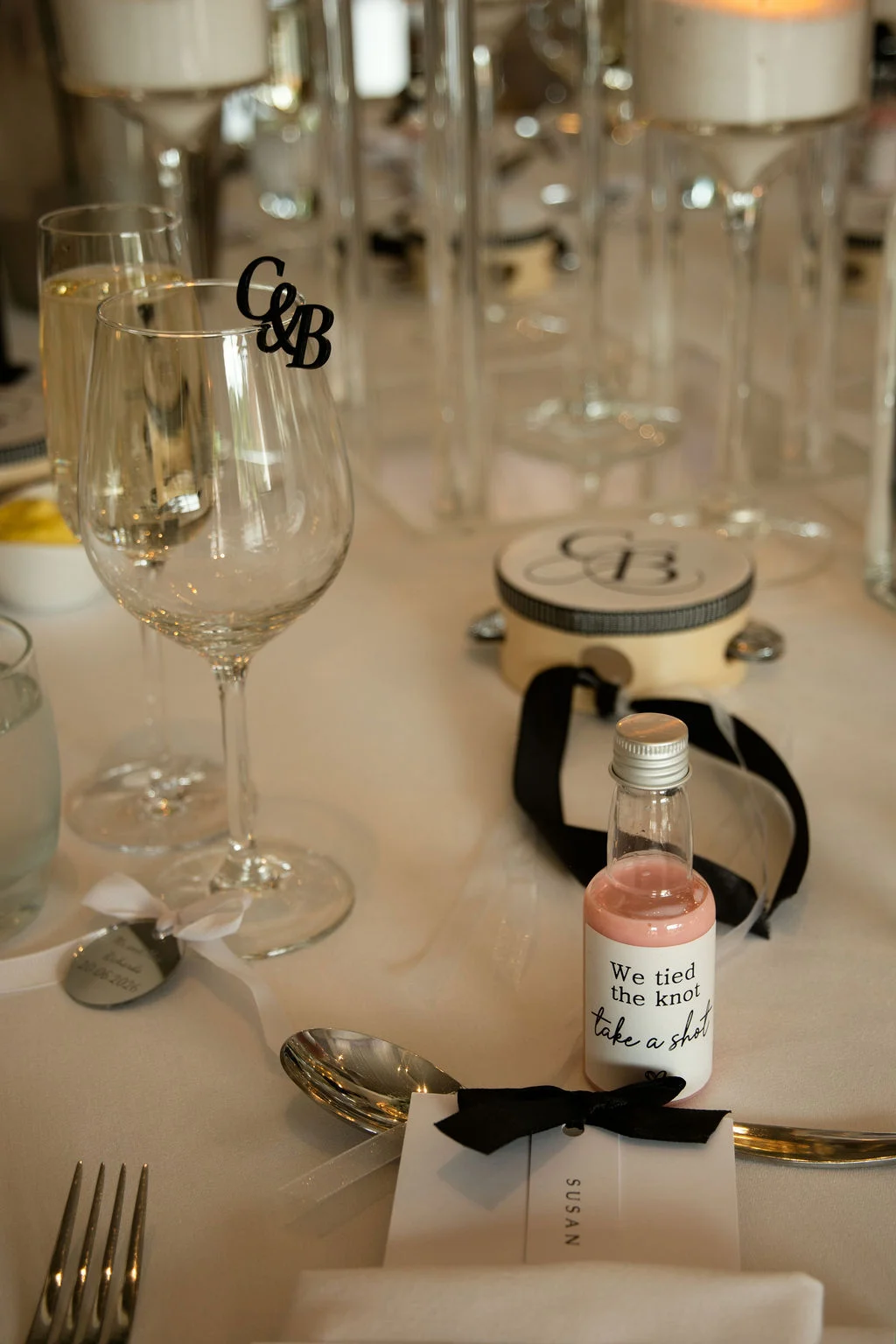 A detailed tablescape features a custom place setting with a personalised favour bottle, place card tied with a black bow, crystal glassware, and a custom monogram glass charm.