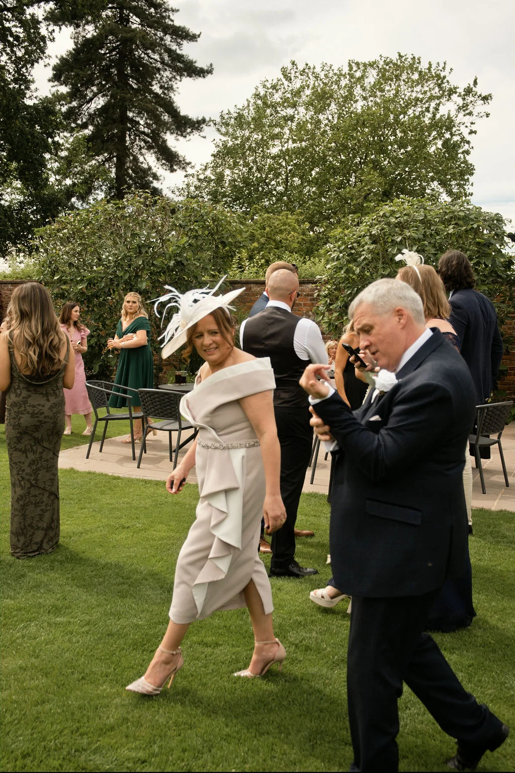 Wedding guests mingle on a lawn during a garden reception, featuring a woman in a chic neutral dress and fascinator walking alongside a man in a dark suit.