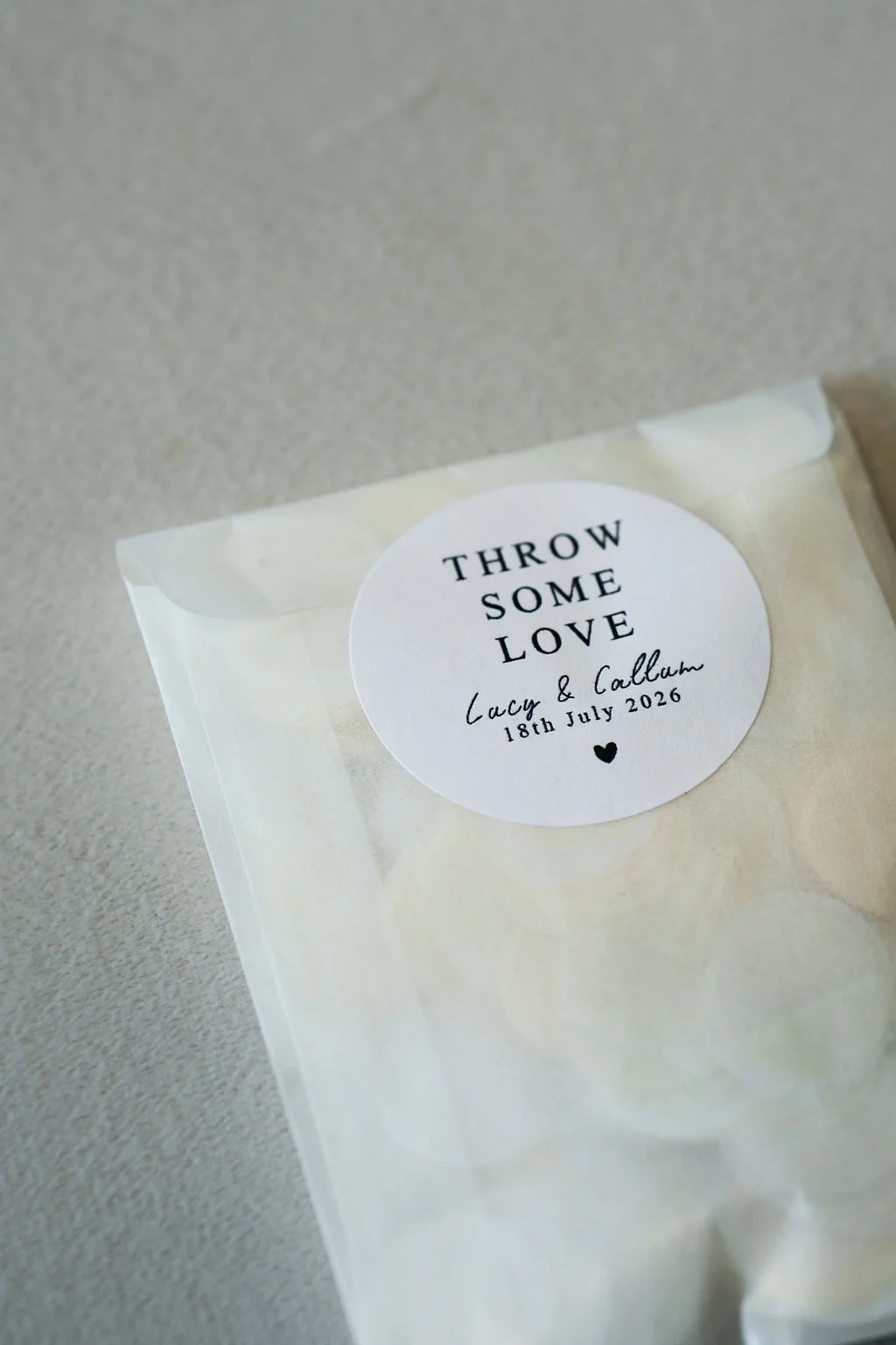 A close-up shot of a translucent glassine wedding confetti packet featuring a round personalised sticker with text reading throw some love.
