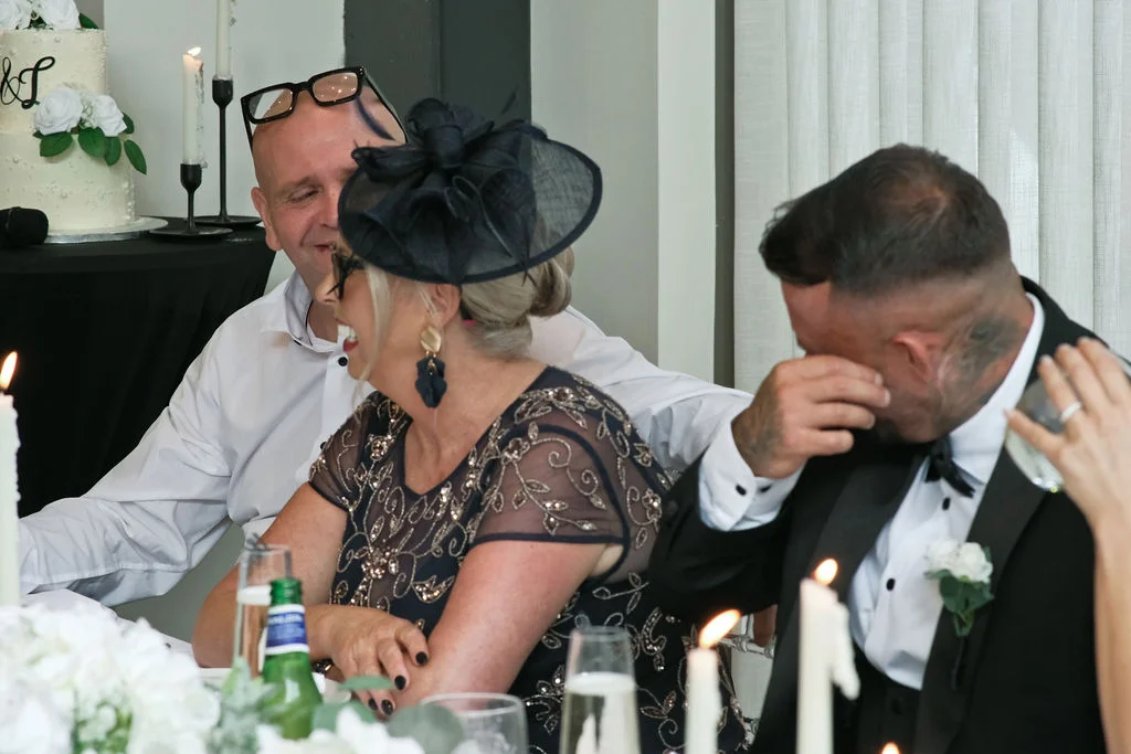 An emotional groom wipes away a tear at the top table during wedding speeches while guests laugh and enjoy the reception.