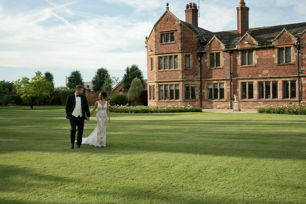 A newlywed couple walks hand in hand across a vast green lawn in front of a grand red brick manor house under a soft blue sky.