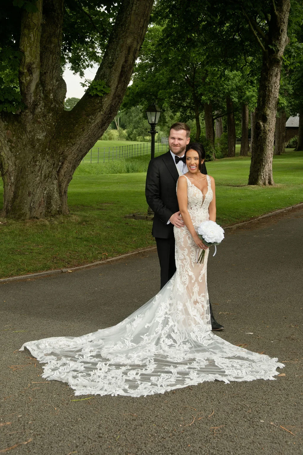 A newlywed couple embraces on a tree-lined driveway, with the laughing bride holding a white rose bouquet and wearing a dramatic lace gown with a long train.