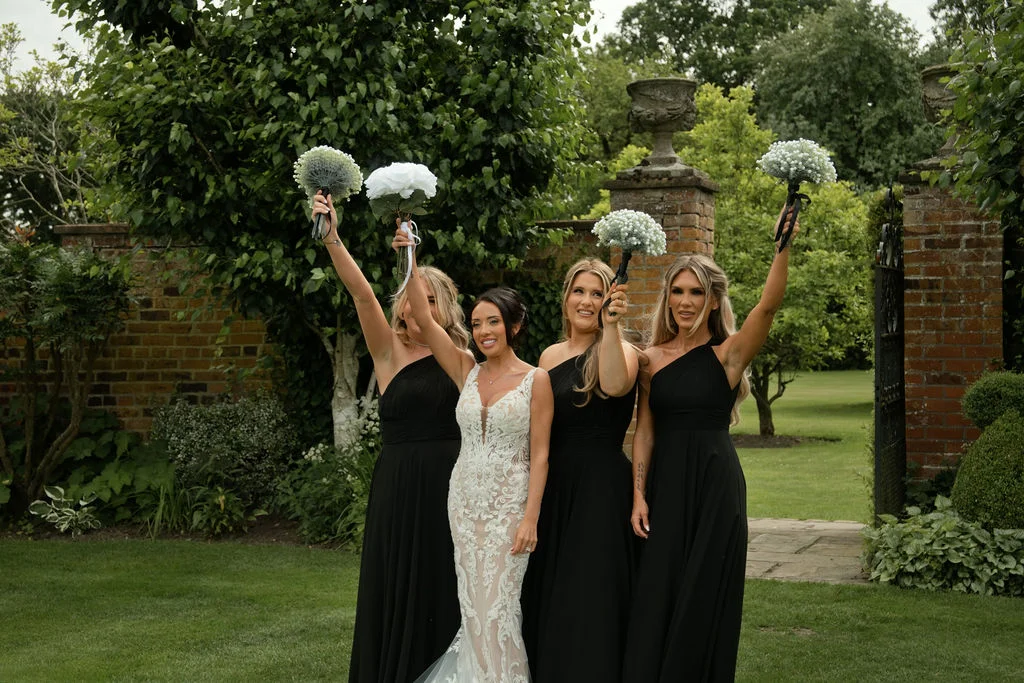 A smiling bride in a detailed lace gown stands in a lush garden flanked by four bridesmaids wearing one-shoulder black dresses holding bouquets high in the air.