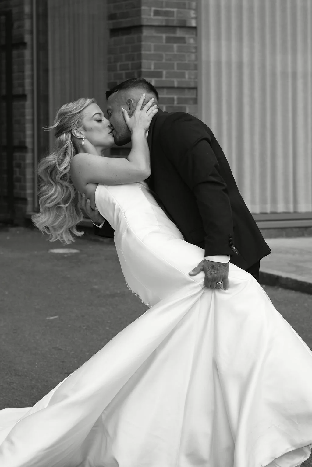 A newlywed couple shares a dramatic dip kiss in front of a brick wall, with the bride wearing a strapless satin gown and her hair in a half-up style.