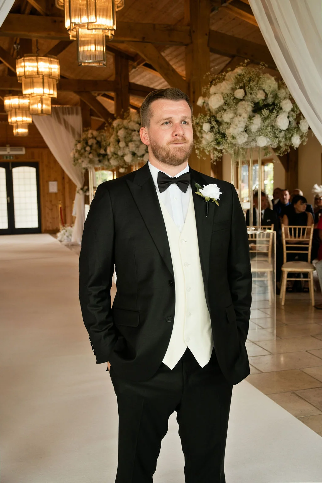 A handsome groom in a black suit, cream waistcoat, and black bow tie waits at the altar inside a rustic oak-framed barn decorated with white floral arrangements.