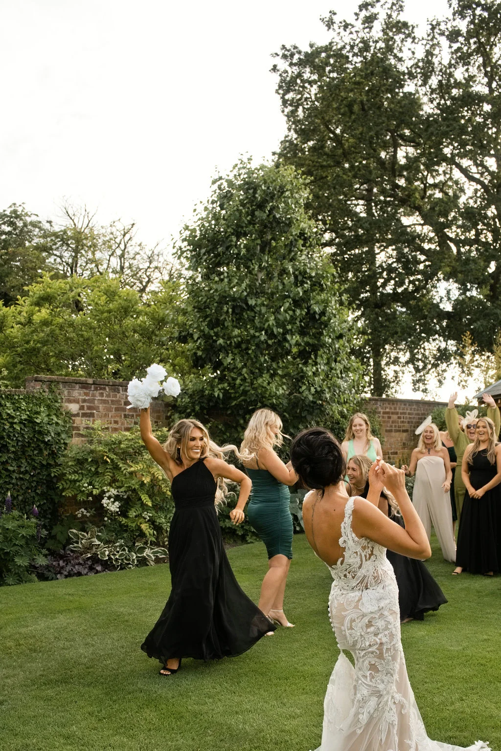 A bride turns away to throw her bouquet on a lush green lawn surrounded by joyful bridesmaids in mismatched dresses.