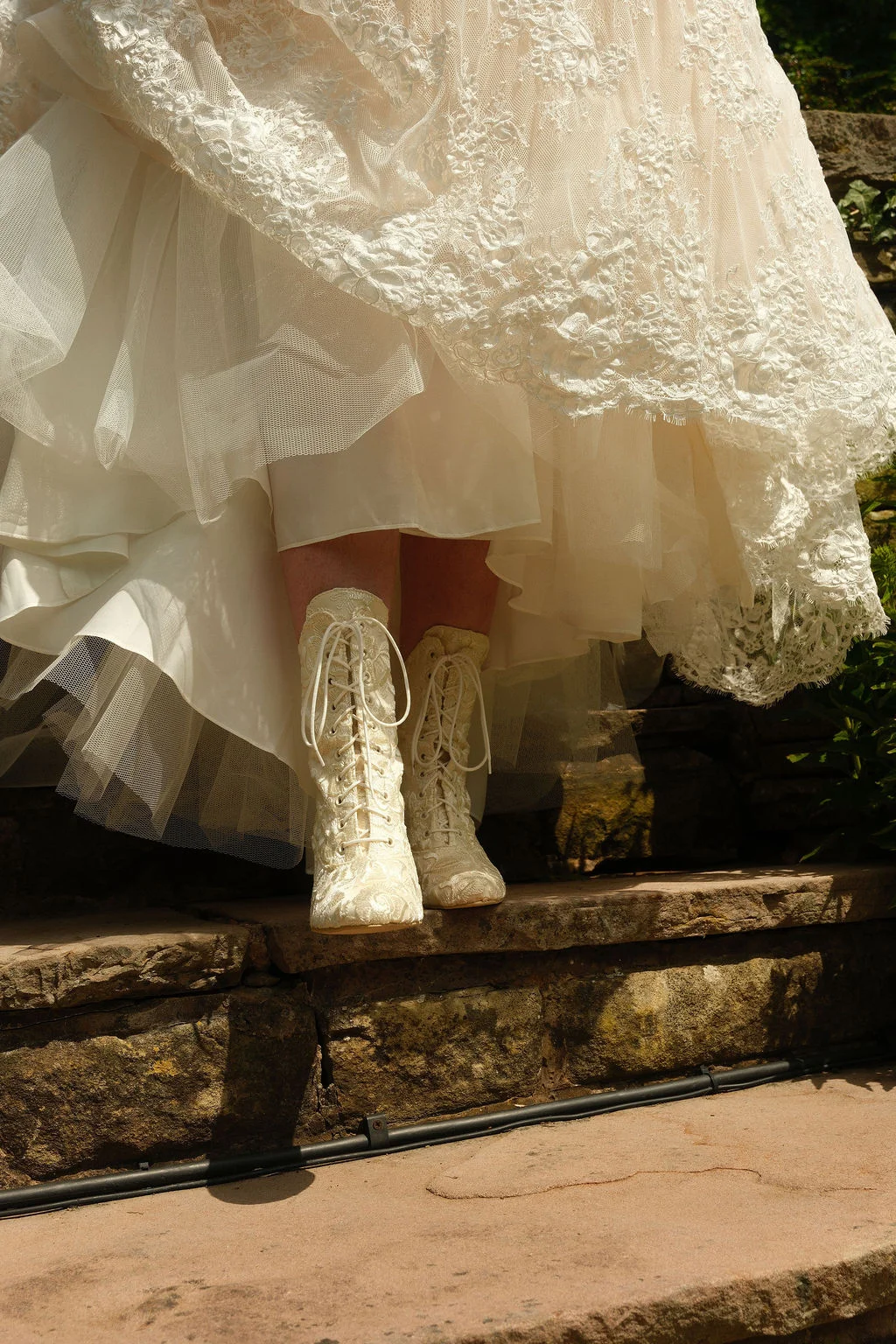 A low-angle shot captures a bride's ivory lace-up wedding boots beneath the layers of a textured lace and tulle gown on rustic stone steps.