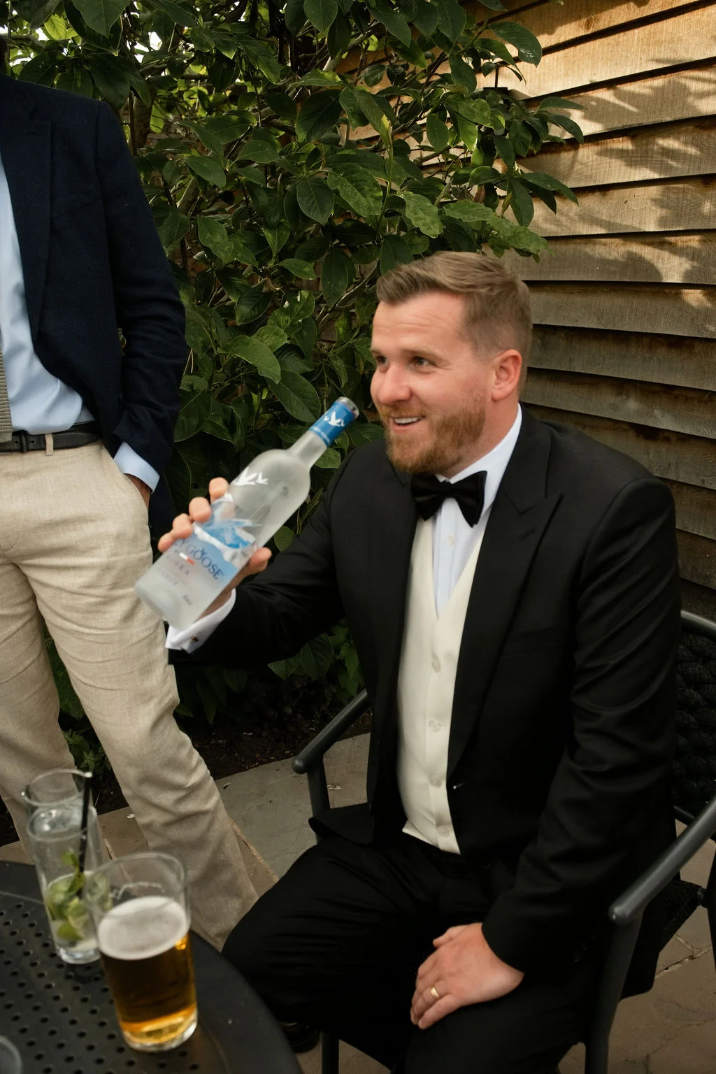 A well-dressed groom wearing a black tuxedo and waistcoat sits outdoors holding a bottle of vodka during the wedding drinks reception.