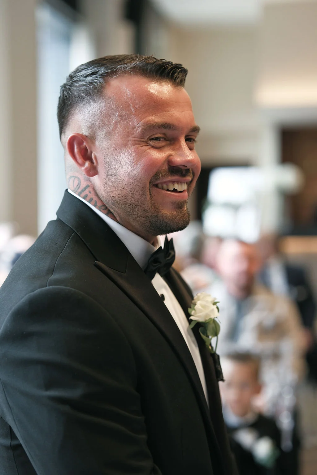 A smiling groom wearing a black tuxedo with a white boutonniere awaits at the altar during the wedding ceremony.