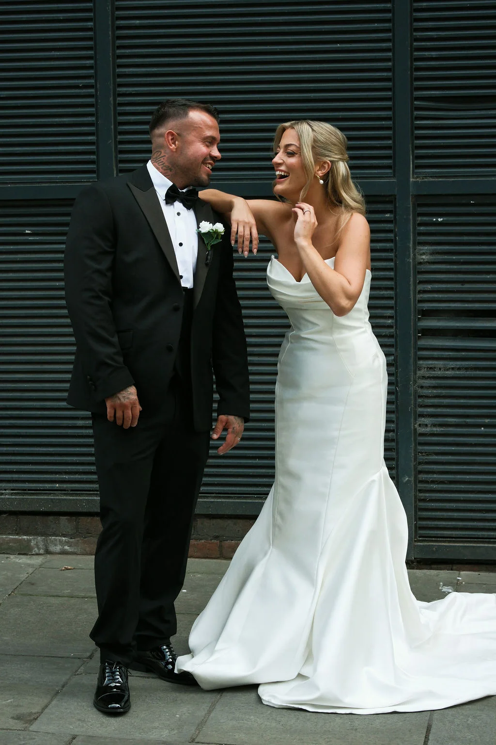 A newlywed couple shares a joyful laugh outside against a dark slatted backdrop, with the bride in a strapless satin trumpet gown and the groom in a classic black tuxedo.