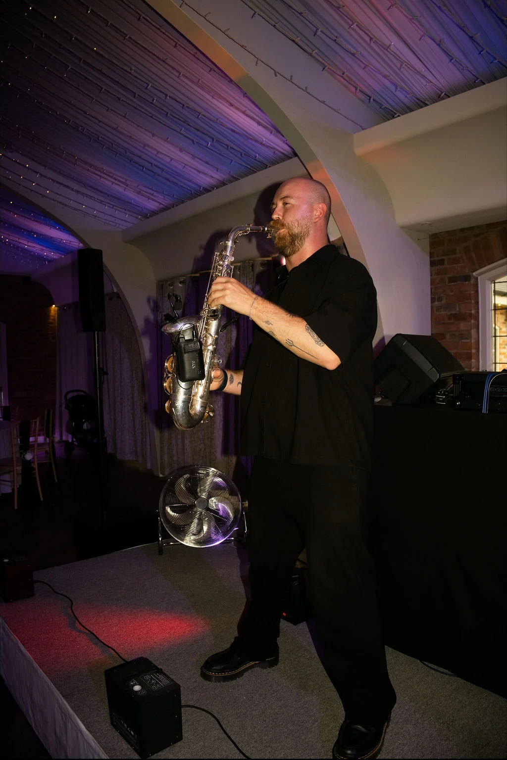 A saxophonist performs on a stage beneath a vaulted ceiling with purple ambient stage lighting during a wedding reception.