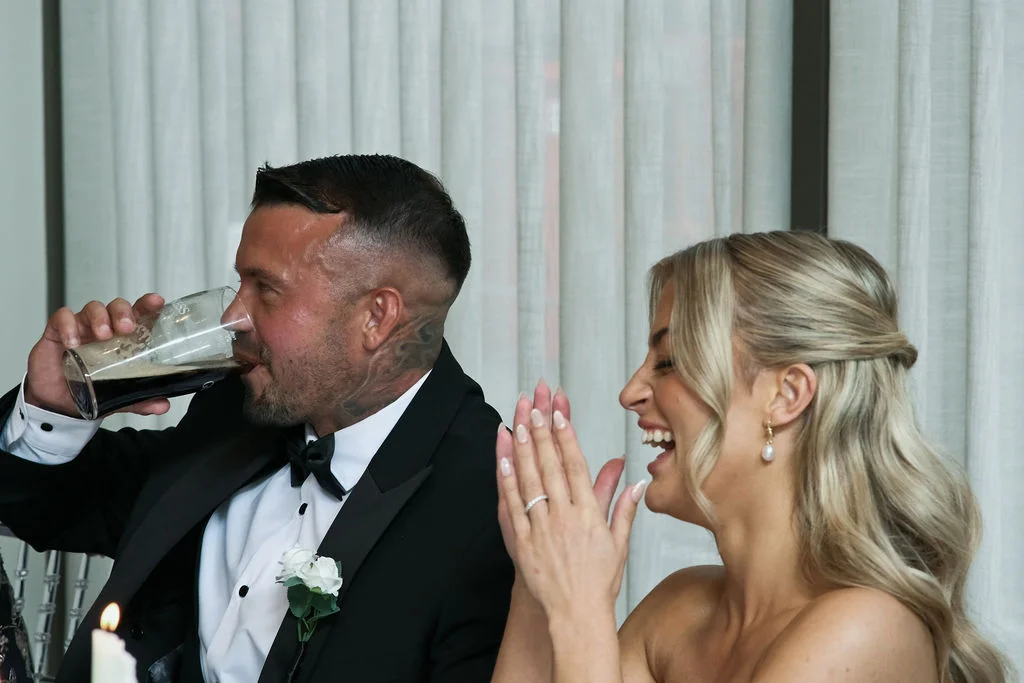 A groom in a classic black tuxedo drinks from a glass while the bride laughs joyfully during the wedding reception speeches.