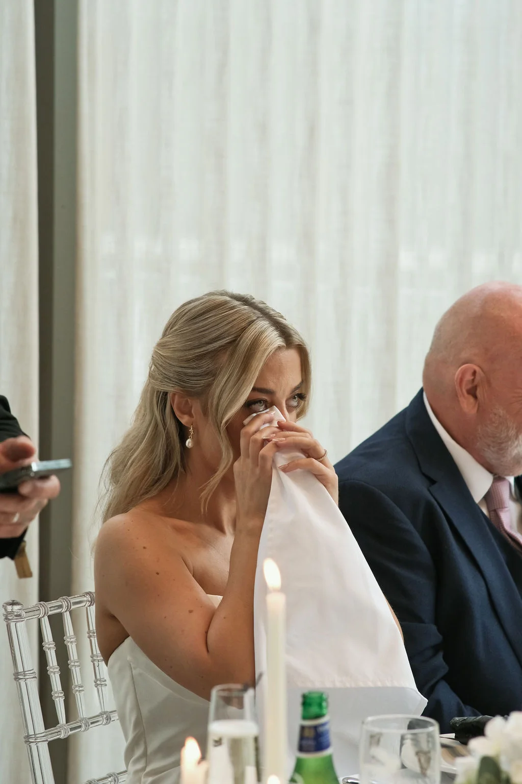An emotional bride gently wipes away tears with a white napkin during the wedding reception speeches, seated beside a guest in a navy suit.