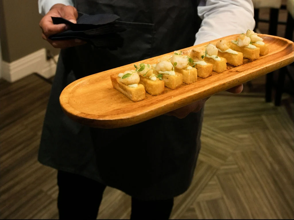 A catering staff member carries a wooden platter lined with gourmet canapes during a wedding reception.