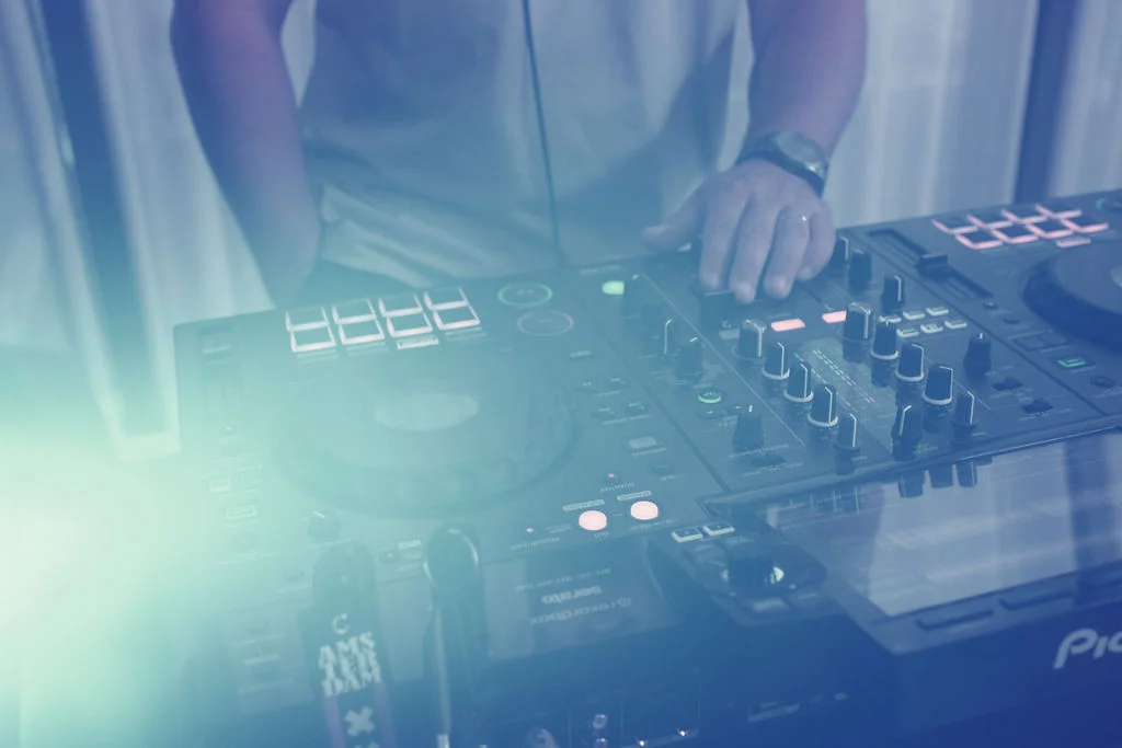 A wedding DJ is performing at the decks, illuminated by atmospheric lighting and cool-toned effects during the evening reception.